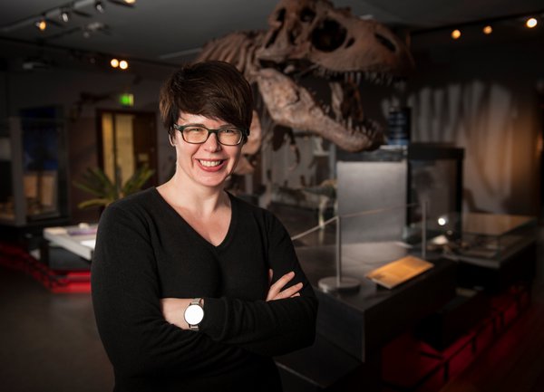 Portrait of Caroline McDonald, stood in front of the replica T. rex skeleton at the Great North Museum