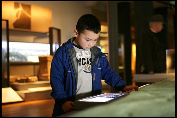 A young boy wearing a Newcastle United FC t-shirt looks at an interactive touchscreen at the Great North Museum